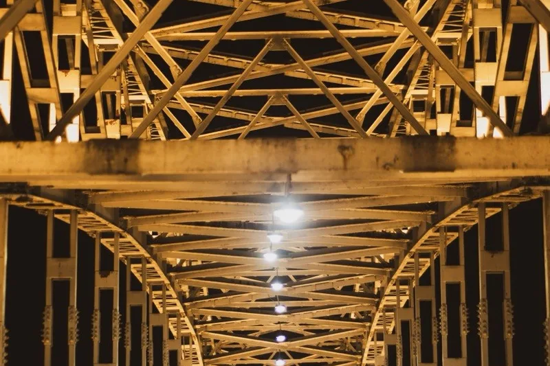 The lit steel truss structure of a bridge, seen from below.
