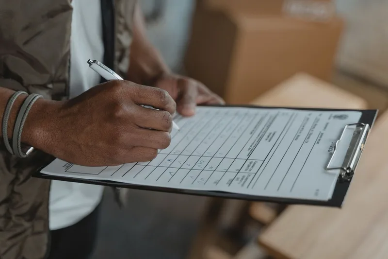 A hand completing a checklist on a clipboard beside packed boxes.